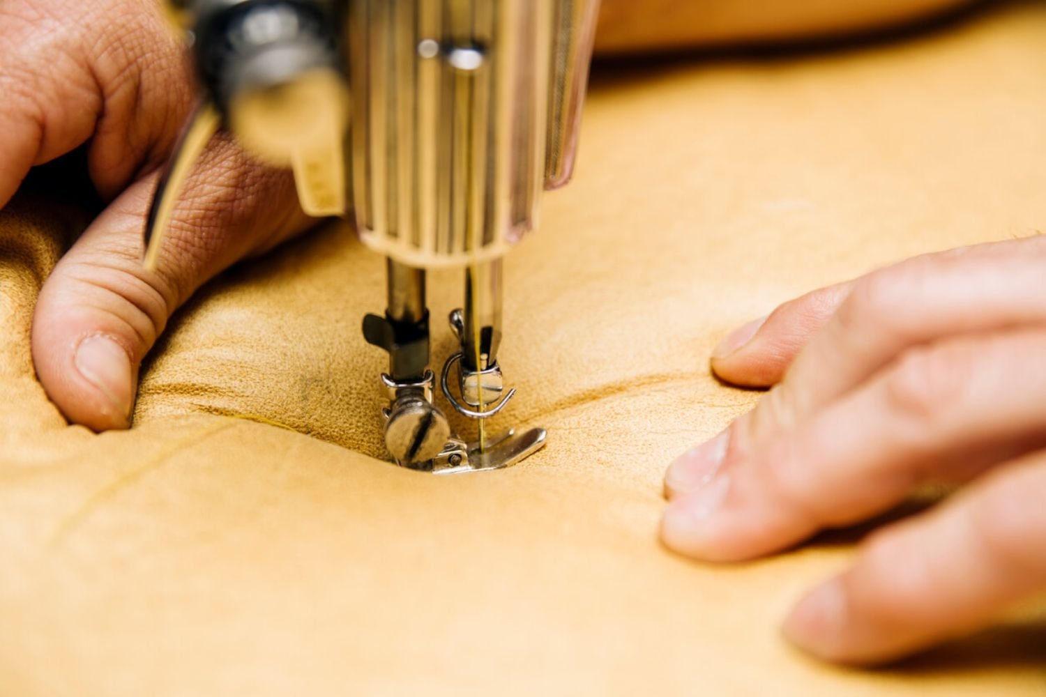 Close-up of hands using a sewing machine to stitch tan leather fabric, demonstrating focused effort on upholstery work.