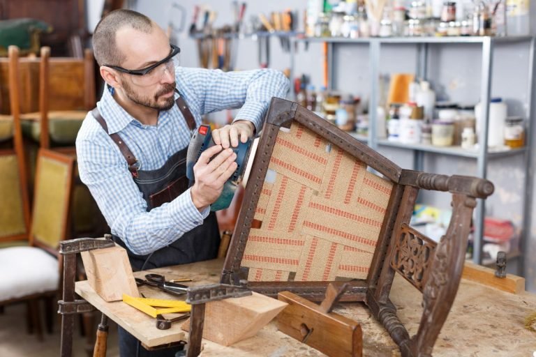 A man is focused on repairing a wooden chair in a well-lit workshop filled with tools and materials.