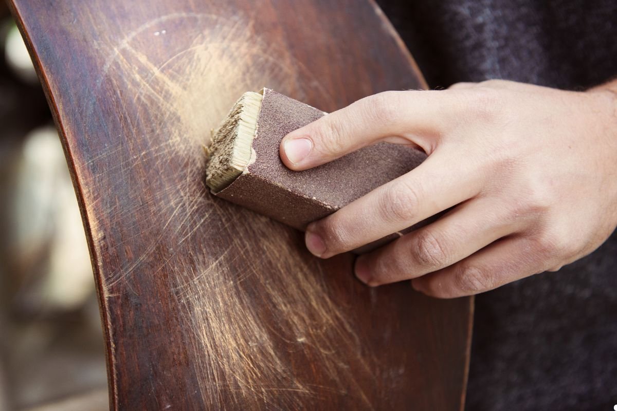 An individual sanding a wooden chair, focusing on refining its surface in a well-lit workspace. 
